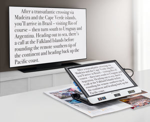 Visolux Digital XL FHD, digital magnifier with large screen, black outer casing and black selection buttons along the bottom of screen, plugged into and transmitting image onto television screen.